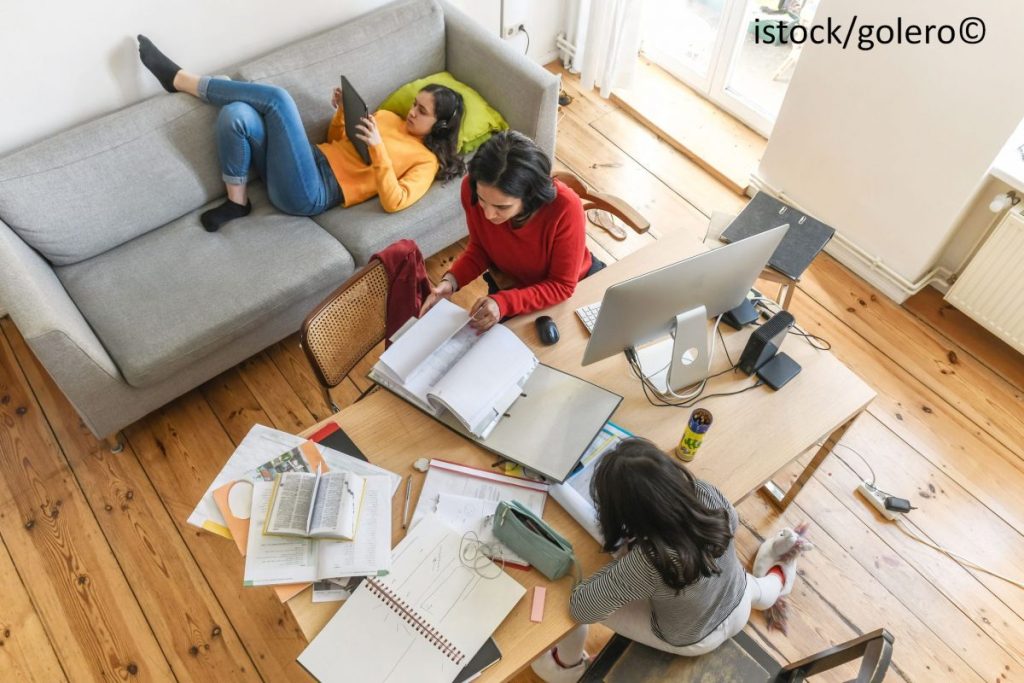 Eine Frau arbeitet am Computer. Gegenüber am Schreibtisch sitzt ein Mädchen und macht Hausaufgaben. Im Hintergrund liegt eine Jugendliche auf der Couch und schaut in ihr Handy.